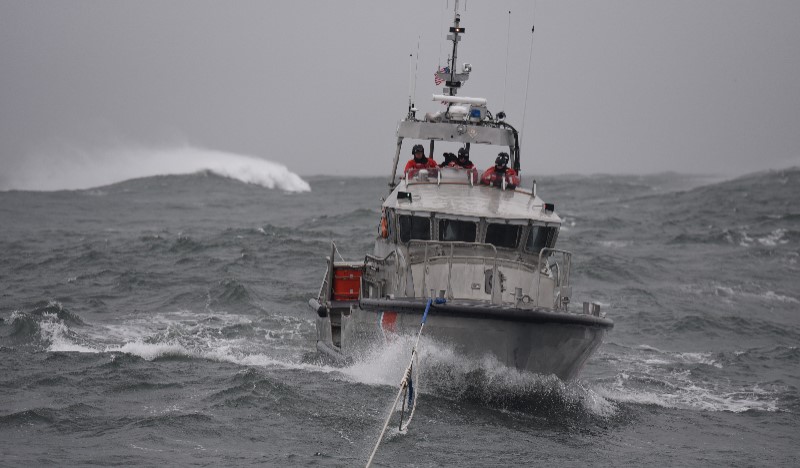 Several coast guard members in orange dry suits and black helmets pilot a silver cutter boat with a rope line attached through rough seas.